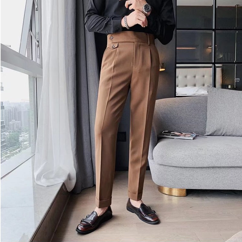 Pantaloni eleganti marroni da uomo, abbinati a mocassini neri, in un ambiente moderno con divano grigio e finestra panoramica.