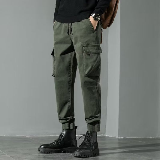 Pantaloni cargo verde militare da uomo, tasche multiple, abbinati a stivali neri e maglione scuro. Stile casual urbano, moda maschile autunnale.