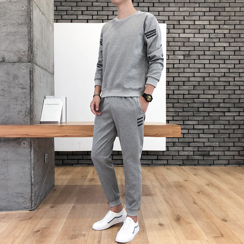 Uomo in tuta sportiva grigia, design moderno, posa in interni minimalisti con parete in mattoni e pavimento in legno, indossa scarpe bianche.