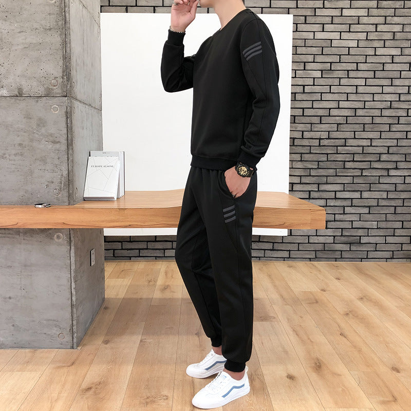 Uomo in tuta sportiva nera con dettagli a righe, posa in un ambiente moderno con pareti in mattoni e pavimento in legno, indossa scarpe bianche.