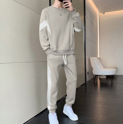 Uomo in tuta sportiva beige con dettagli bianchi, scarpe da ginnastica bianche, in un interno moderno con pareti chiare e pavimento scuro.