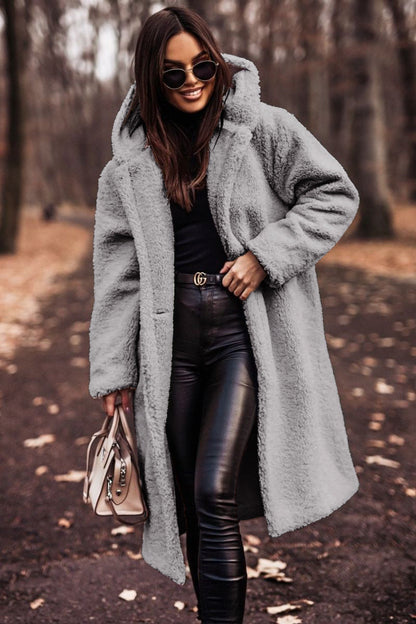 Donna con cappotto teddy grigio, occhiali da sole, pantaloni in pelle nera, borsa beige, cammina in un parco autunnale. Moda autunno inverno.