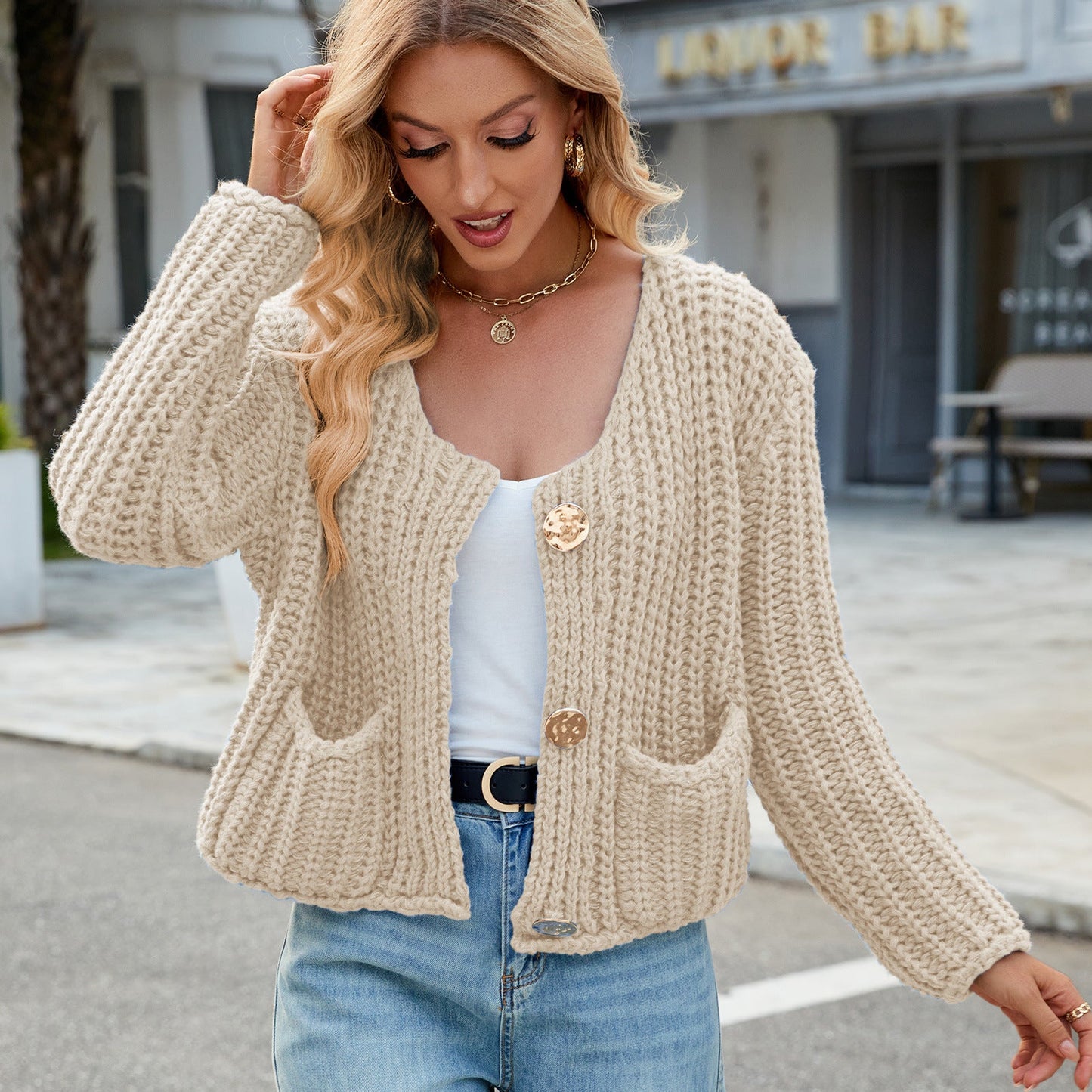 Donna con cardigan beige lavorato a maglia, indossa jeans e top bianco. Moda autunnale casual, strada cittadina sullo sfondo.