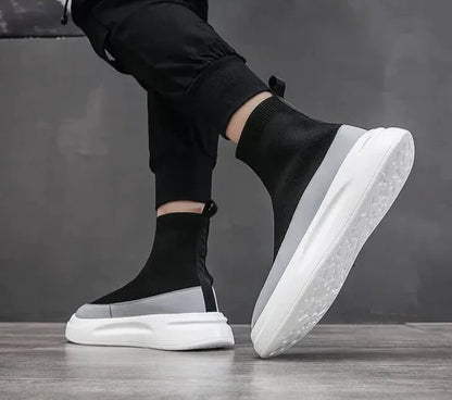 Scarpe da ginnastica nere e grigie con suola bianca, design moderno e minimalista, indossate da una persona su pavimento grigio.