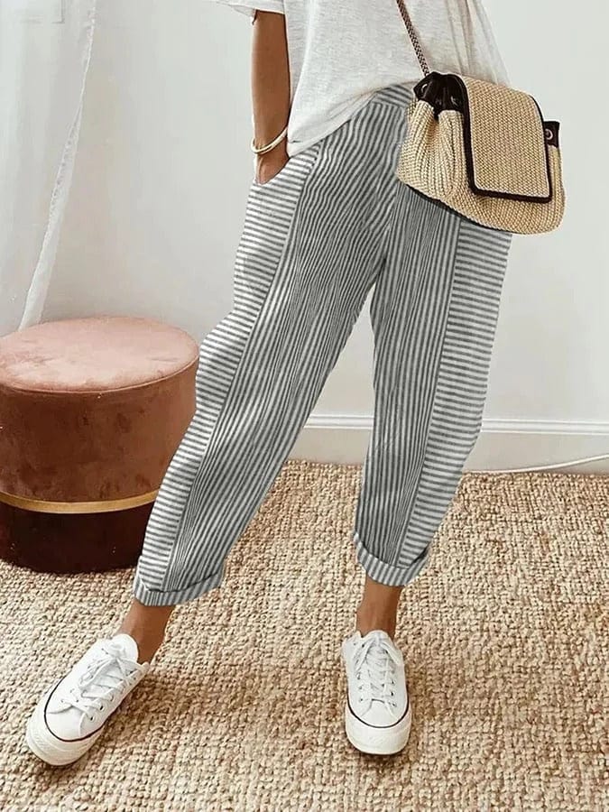 Donna con pantaloni a righe bianche e grigie, maglietta bianca, borsa a tracolla in paglia, scarpe da ginnastica bianche su tappeto beige.