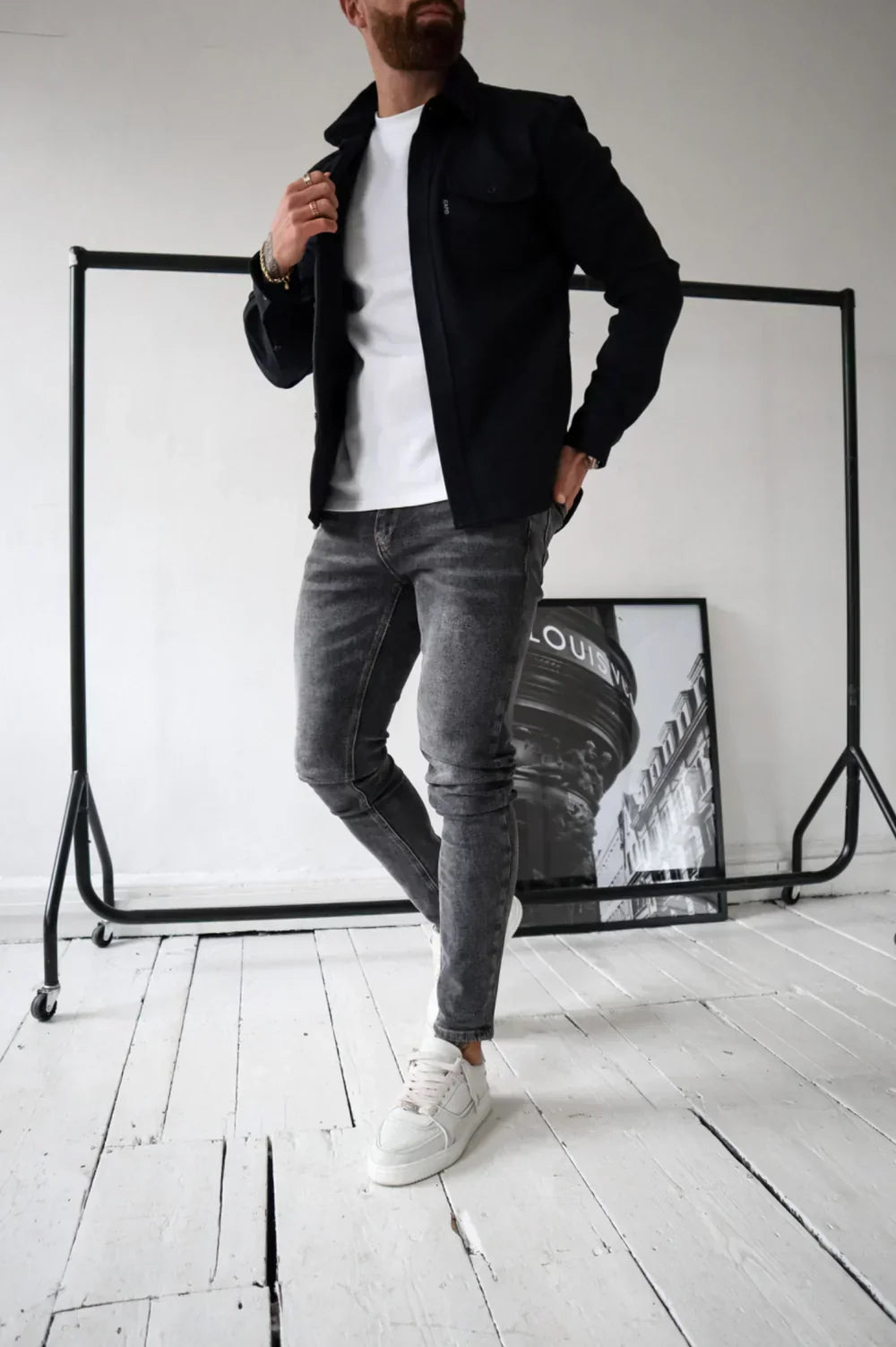 Uomo con giacca nera, jeans grigi e scarpe bianche su pavimento in legno bianco, sfondo con cornice fotografica. Moda casual maschile.