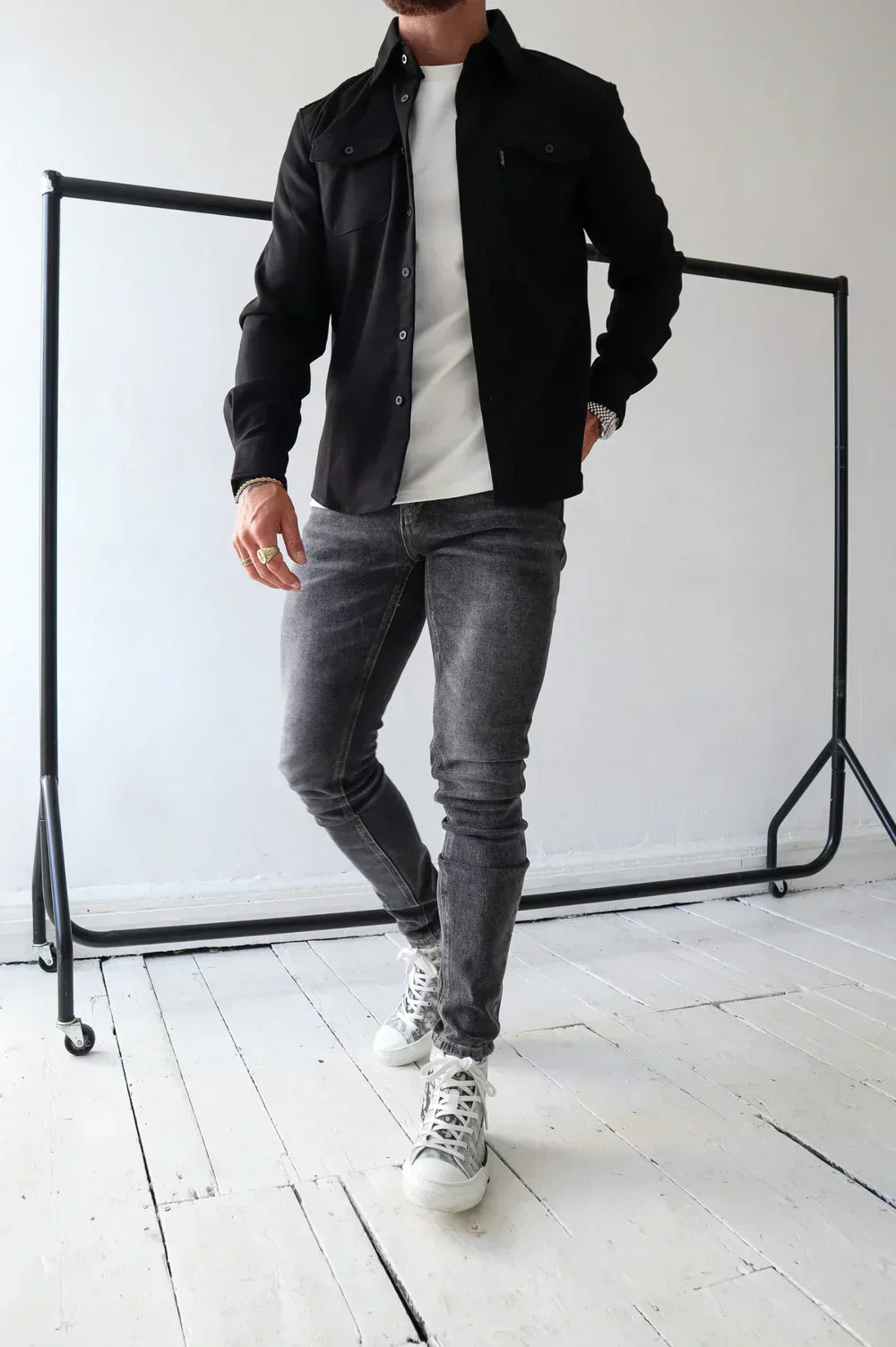 Uomo in giacca nera e jeans grigi, indossa sneakers bianche, posa su pavimento in legno bianco. Moda casual maschile, abbigliamento moderno.