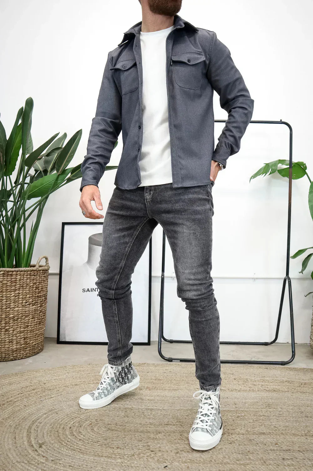 Uomo in jeans grigi e giacca blu, con sneakers bianche, in posa moderna. Sfondo con piante e decorazioni minimaliste. Moda casual maschile.