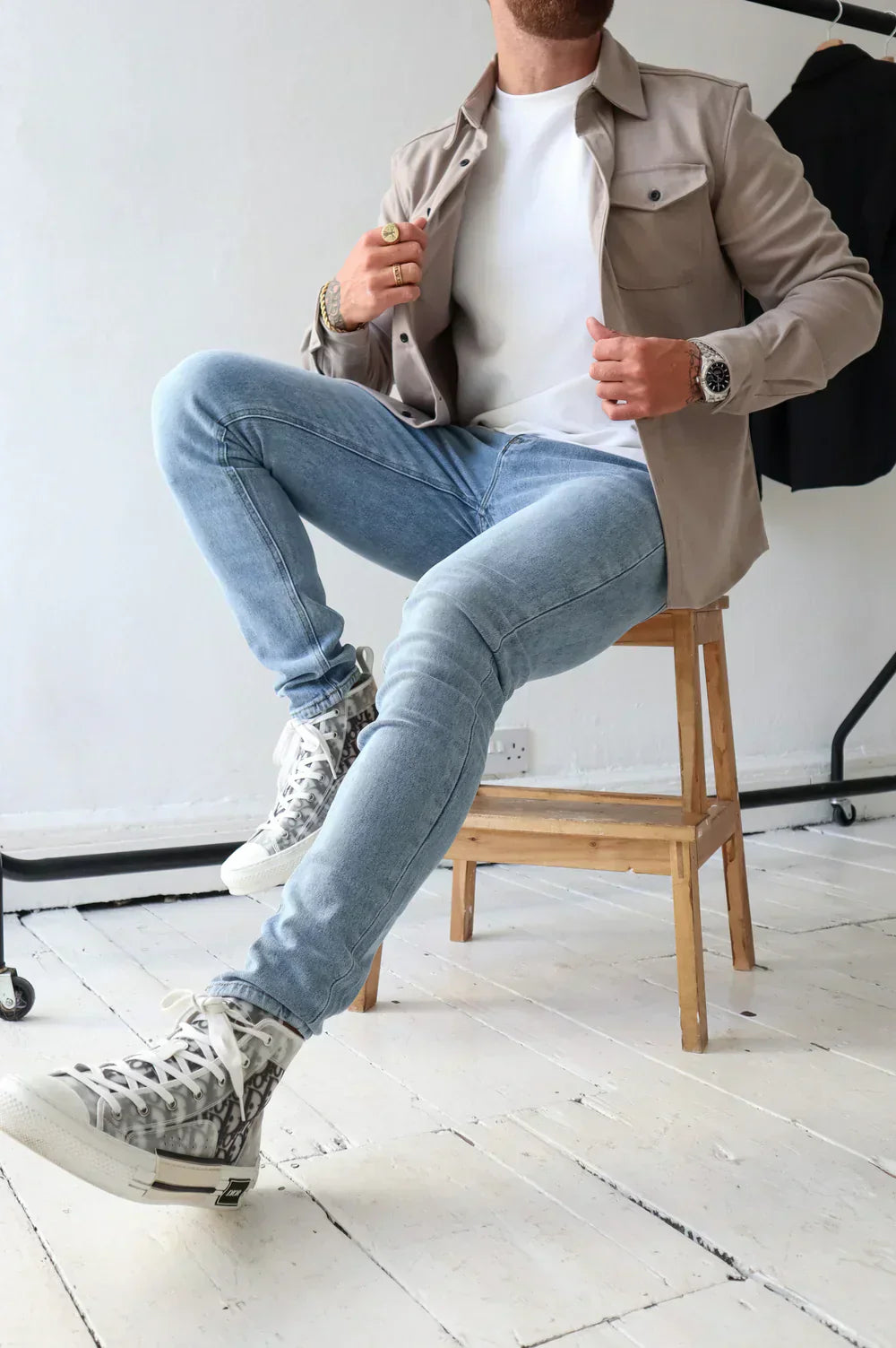Uomo seduto su sedia indossa jeans chiari, maglietta bianca, giacca beige e sneakers alte. Moda casual maschile, stile urbano moderno.