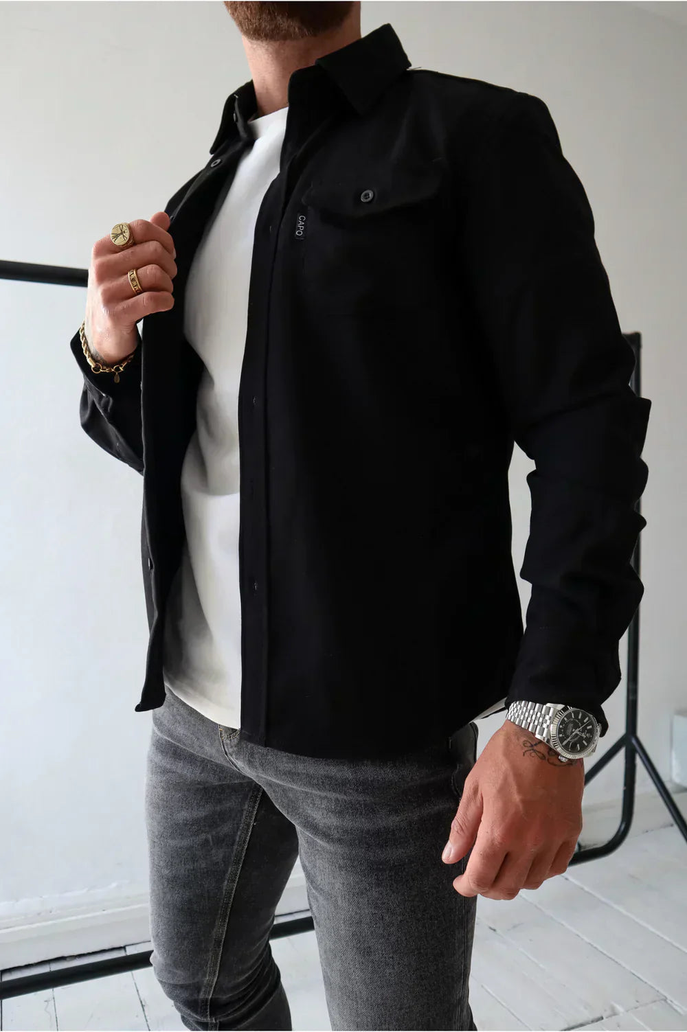 Uomo con camicia nera aperta su t-shirt bianca, jeans grigi, anelli dorati e orologio argento. Moda maschile casual elegante.