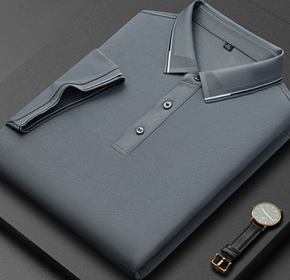 Camicia polo grigia piegata su sfondo nero, con colletto a contrasto e bottoni, accanto a un orologio elegante. Moda uomo, abbigliamento casual.