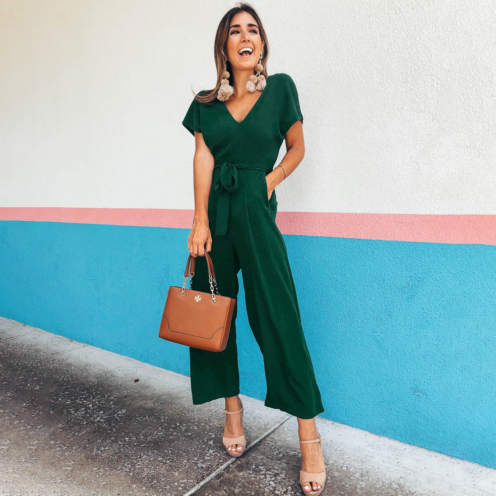 Donna sorridente in tuta verde elegante, con borsa marrone e sandali beige, posa davanti a muro colorato. Moda femminile, abbigliamento estivo chic.