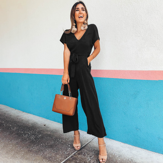 Donna sorridente in tuta nera elegante, con borsa marrone e sandali beige, posa davanti a muro colorato. Moda femminile, stile estivo chic.