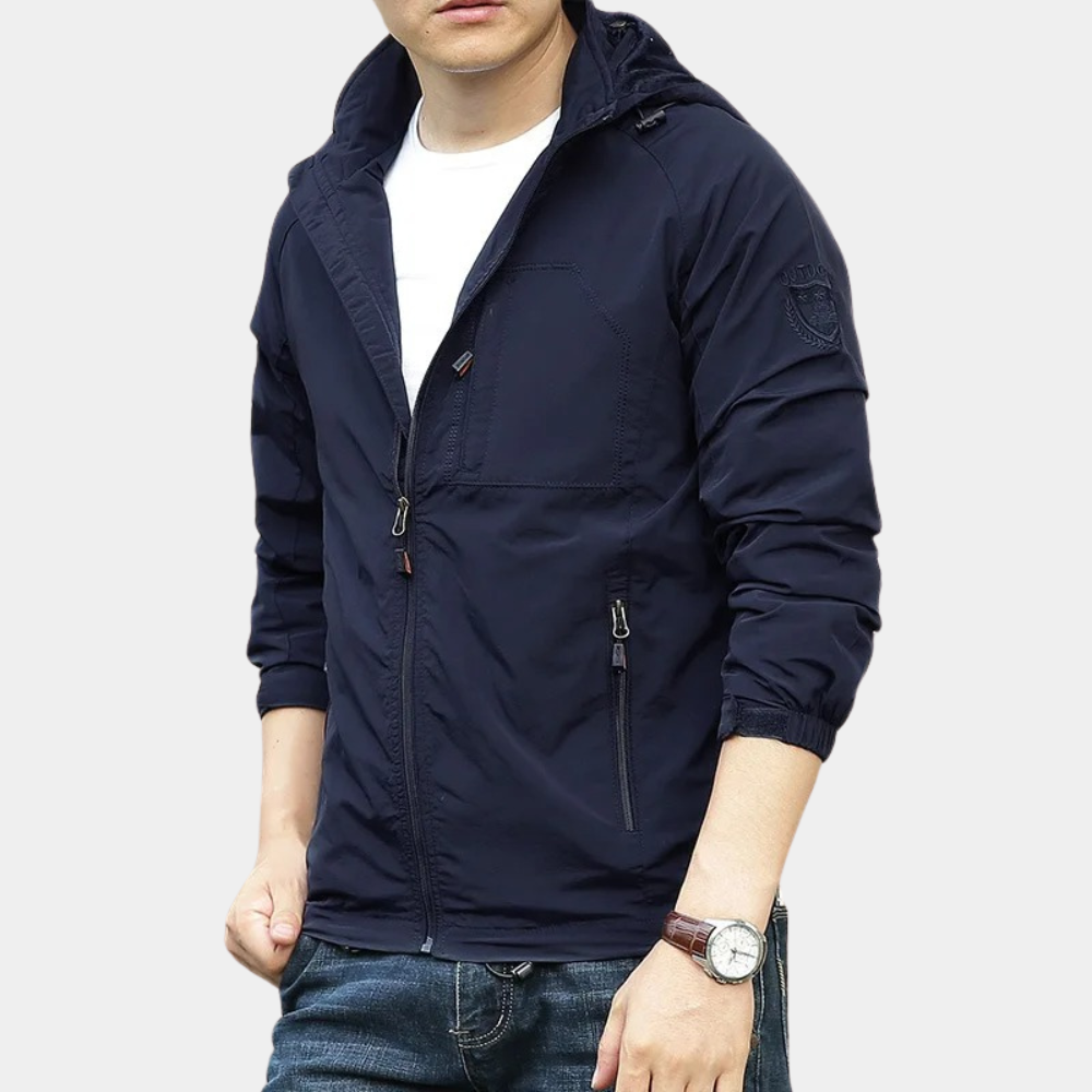 Giacca a vento uomo blu scuro, con cappuccio e cerniere, indossata su maglietta bianca e jeans. Stile casual, moda maschile autunnale.