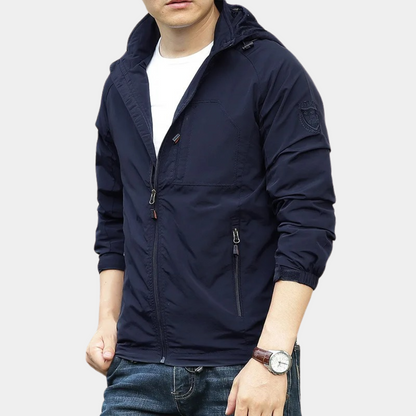 Giacca a vento uomo blu scuro, con cappuccio e cerniere, indossata su maglietta bianca e jeans. Stile casual, moda maschile autunnale.