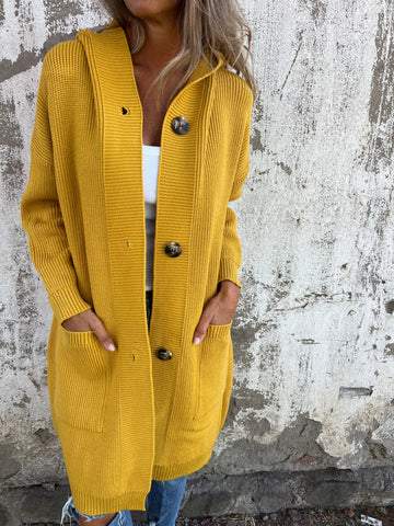 Donna con cardigan giallo senape, lungo e con bottoni grandi, su sfondo di muro rustico. Moda autunnale, abbigliamento casual elegante.
