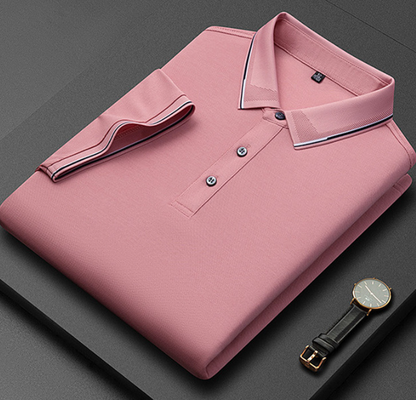 Camicia polo rosa piegata su sfondo grigio, con colletto a contrasto e due bottoni visibili. Accanto, un orologio con cinturino in pelle nera.