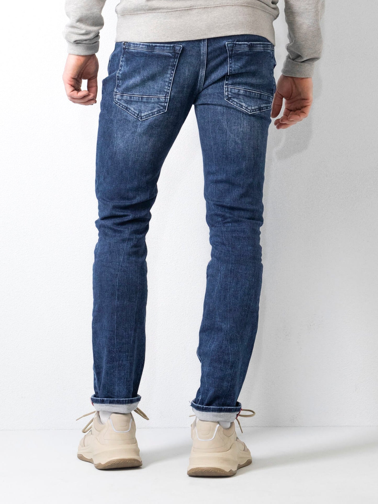 Jeans uomo slim fit blu, visti da dietro, abbinati a scarpe sportive beige e felpa grigia. Moda casual maschile, abbigliamento denim.