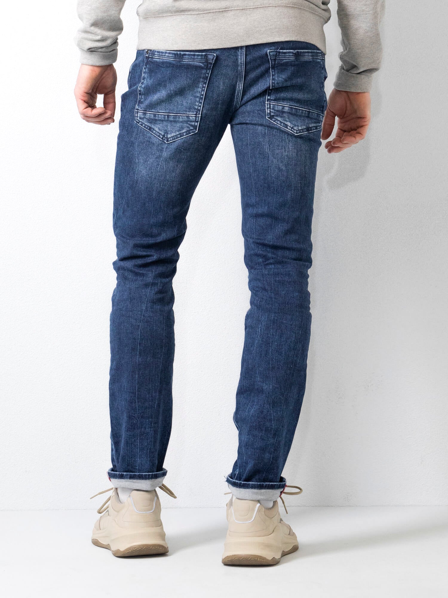 Jeans uomo slim fit blu, visti da dietro, abbinati a scarpe sportive beige e felpa grigia. Moda casual maschile, abbigliamento denim.