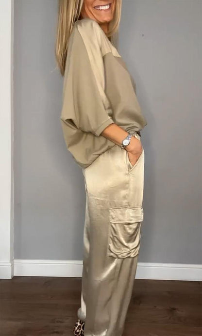 Donna in abbigliamento elegante beige, camicia ampia e pantaloni satinati con tasche, posa sorridente su sfondo grigio. Moda femminile chic.