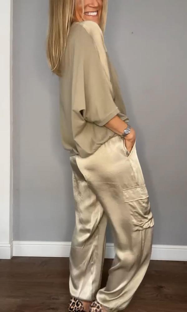Donna in abbigliamento elegante beige, indossa pantaloni e maglia di seta, posa sorridente su sfondo grigio. Moda femminile, stile sofisticato.
