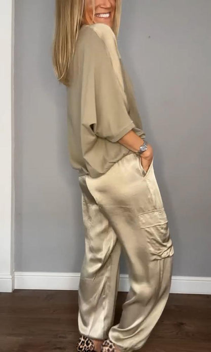 Donna in abbigliamento elegante beige, indossa pantaloni e maglia di seta, posa sorridente su sfondo grigio. Moda femminile, stile sofisticato.