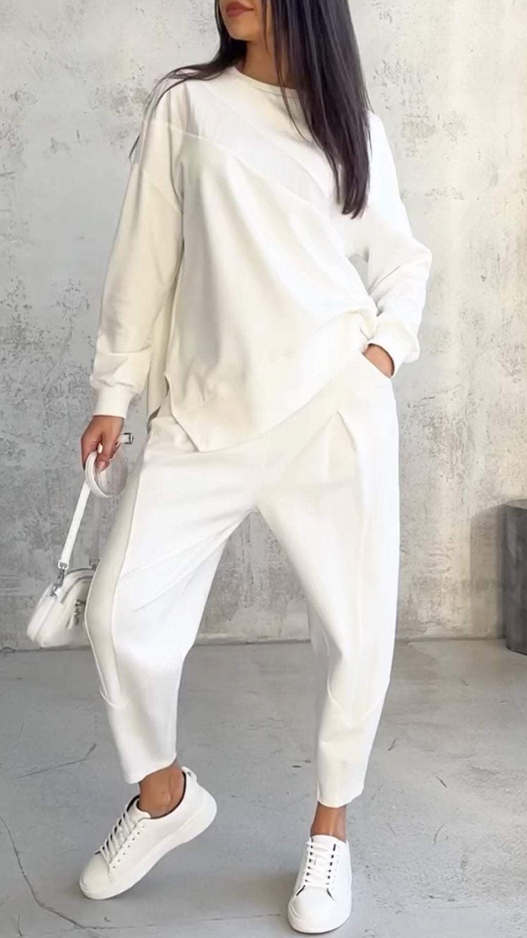 Donna in abbigliamento casual bianco, con felpa e pantaloni coordinati, scarpe da ginnastica bianche e borsa a tracolla, su sfondo grigio minimalista.