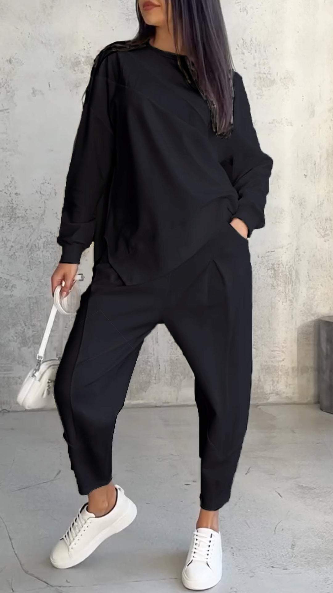 Donna in abbigliamento casual nero, con felpa e pantaloni larghi, borsa bianca e sneakers bianche, su sfondo grigio. Moda urbana femminile.