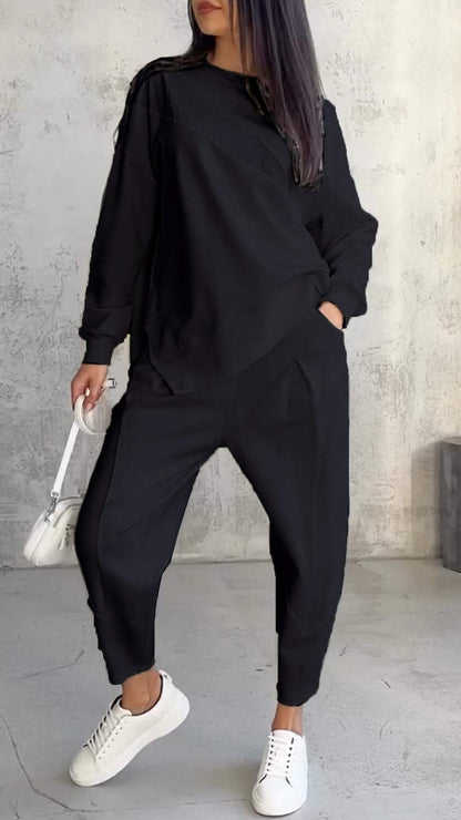 Donna in abbigliamento casual nero, con felpa e pantaloni larghi, borsa bianca e sneakers bianche, su sfondo grigio. Moda urbana femminile.