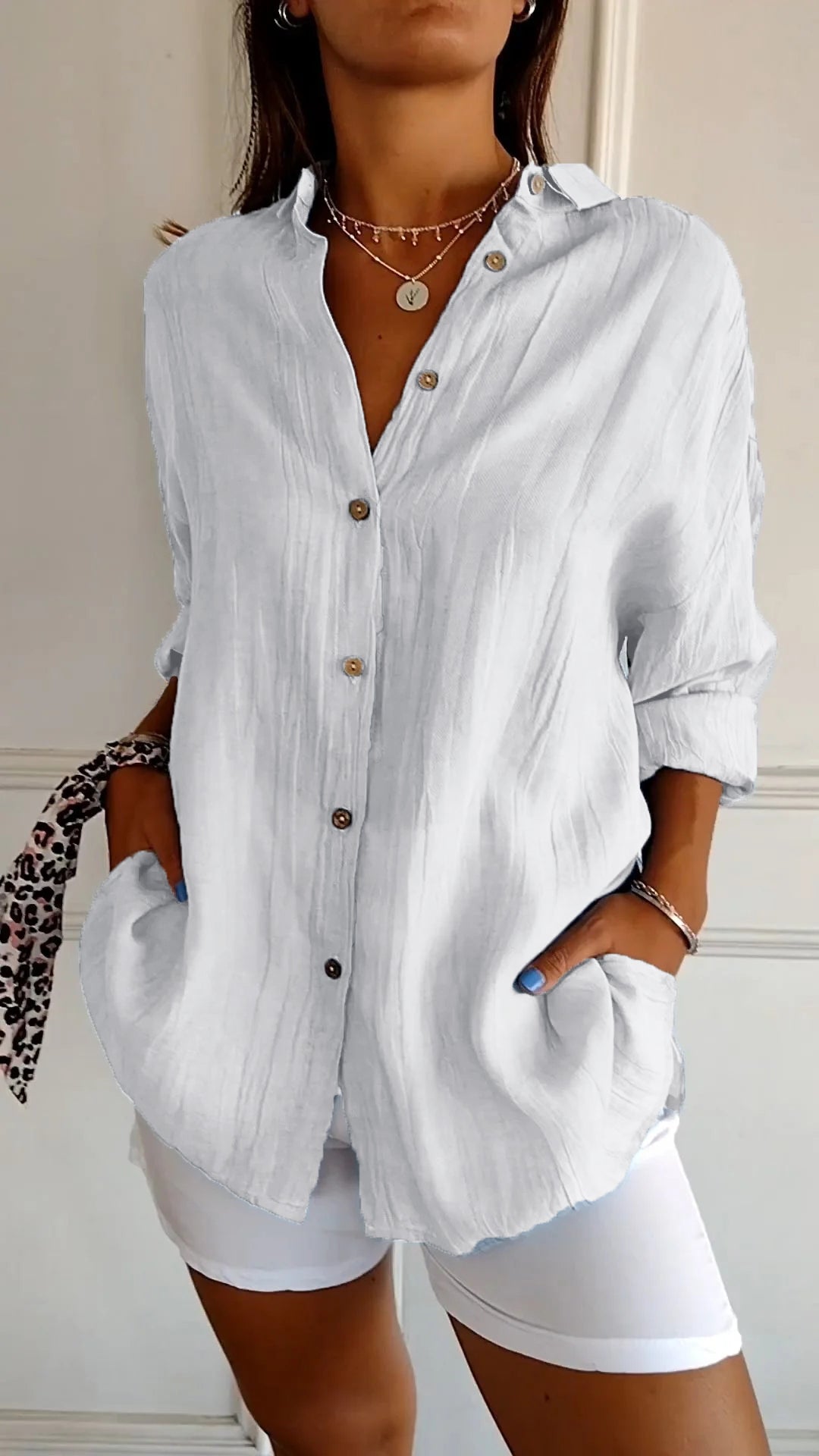 Camicia bianca in lino con bottoni marroni, maniche lunghe arrotolate, indossata con pantaloni bianchi. Stile casual elegante, moda donna.
