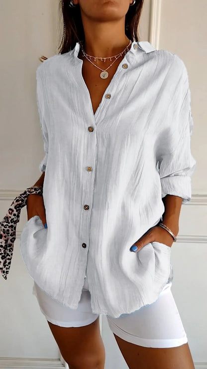 Camicia bianca in lino con bottoni marroni, maniche lunghe arrotolate, indossata con pantaloni bianchi. Stile casual elegante, moda donna.