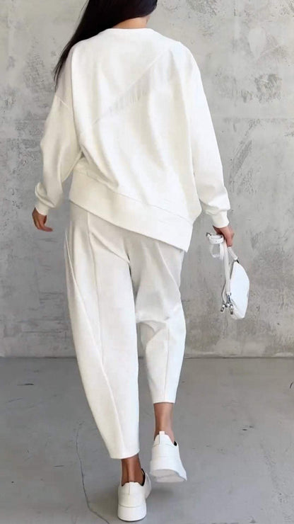 Donna di spalle in tuta bianca elegante, con borsa a mano bianca e sneakers, su sfondo grigio minimalista. Moda casual chic autunnale.