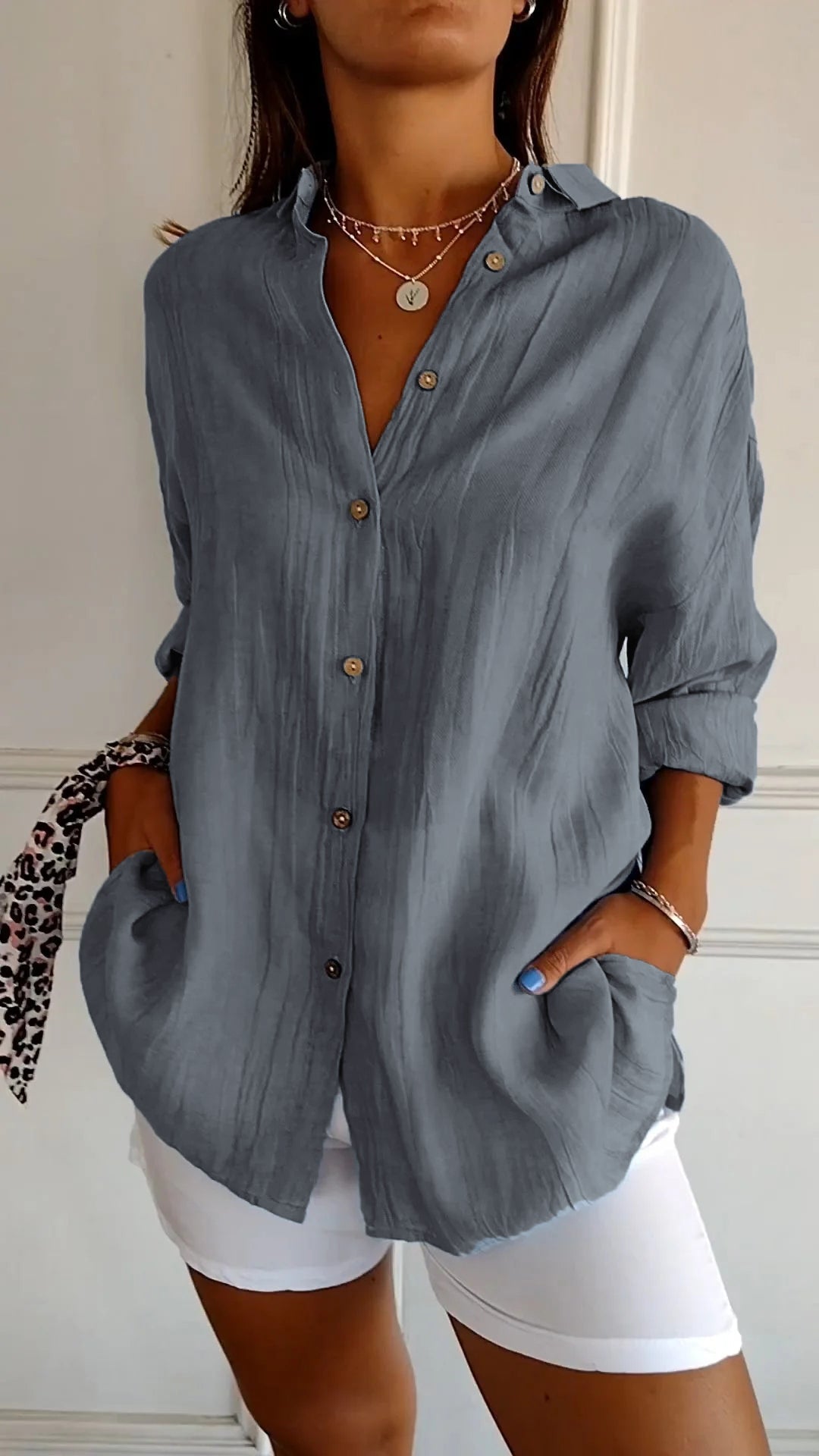Camicia grigia a maniche lunghe con bottoni, indossata con pantaloni bianchi. Stile casual elegante, accessoriata con collana e bracciale.