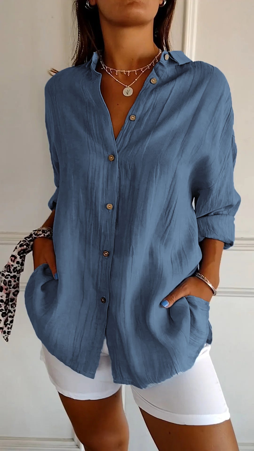 Donna con camicia blu a maniche lunghe, bottoni in legno, tasche frontali, abbinata a pantaloni bianchi. Stile casual elegante, moda femminile.