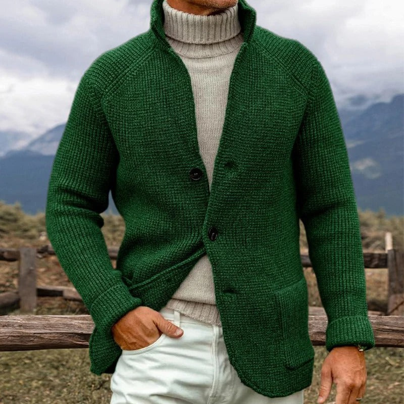 Uomo con cardigan verde a maglia grossa, collo alto, su sfondo montano. Moda autunnale, abbigliamento caldo, stile casual elegante.