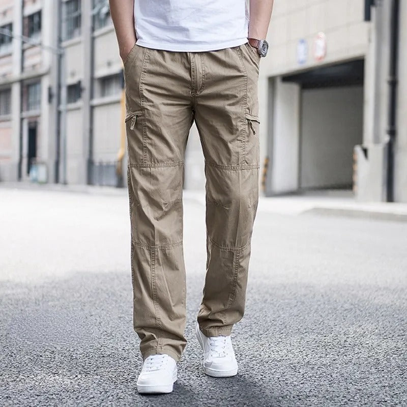 Pantaloni cargo beige da uomo, tasche multiple, indossati con scarpe bianche, sfondo urbano. Moda casual maschile, abbigliamento streetwear.