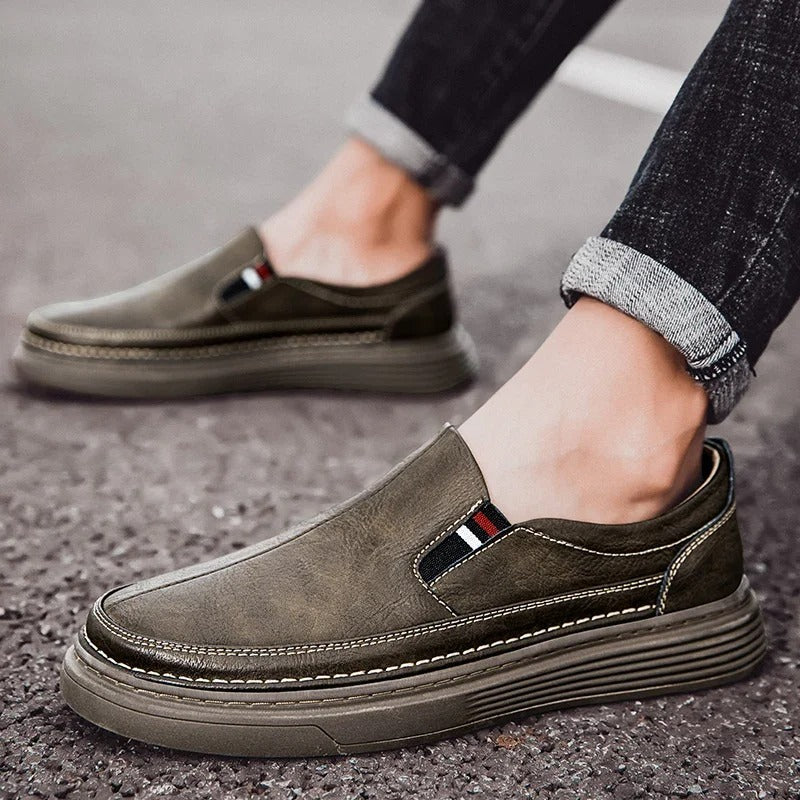 Scarpe slip-on da uomo in pelle marrone, suola spessa, dettagli cuciture bianche, indossate con jeans. Calzature casual eleganti e comode.