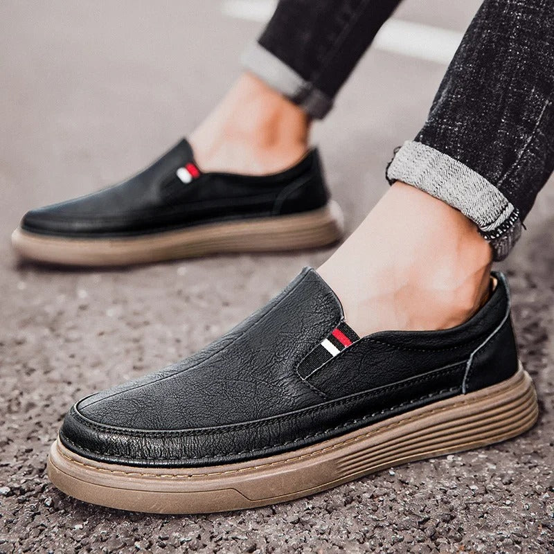 Scarpe slip-on uomo in pelle nera, suola marrone, dettagli a righe rosse e bianche, indossate con jeans. Calzature eleganti e casual.
