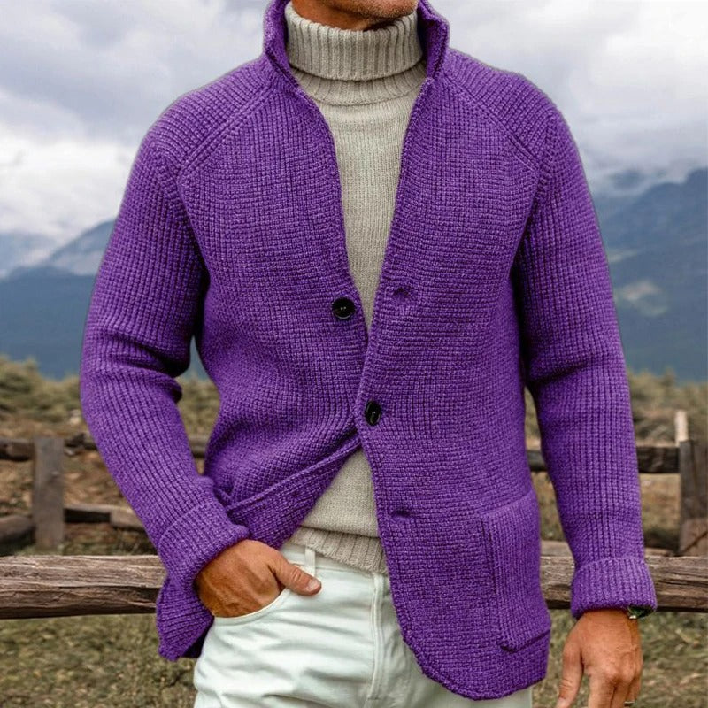 Uomo con maglione viola a maglia grossa e dolcevita beige, sfondo montano. Moda invernale, abbigliamento caldo, stile casual elegante.