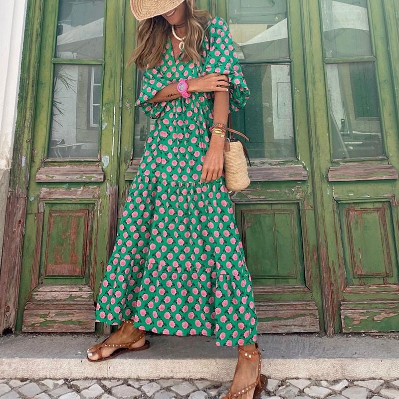 Donna con abito lungo verde a fiori rosa, cappello di paglia, borsa di paglia, sandali marroni davanti a porte verdi antiche. Moda estiva elegante.