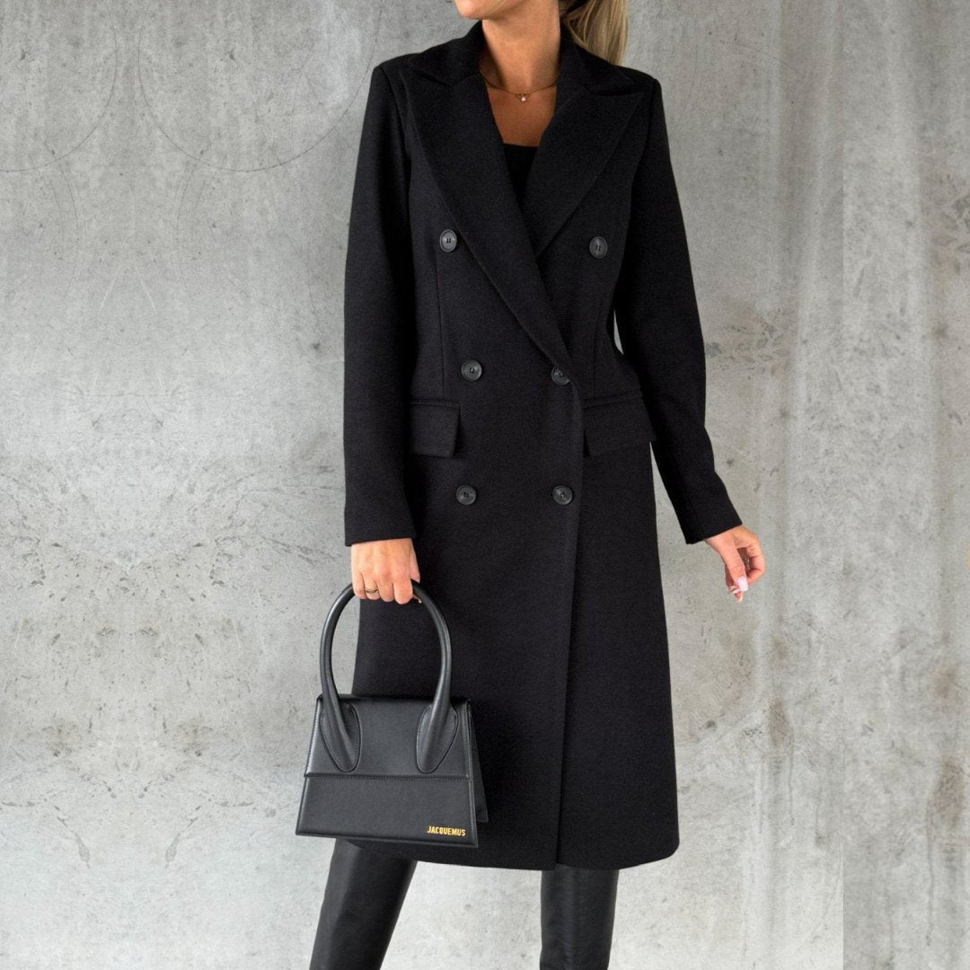 Donna in cappotto nero elegante, doppio petto, con borsa nera, sfondo grigio. Moda autunnale, stile sofisticato, abbigliamento femminile chic.