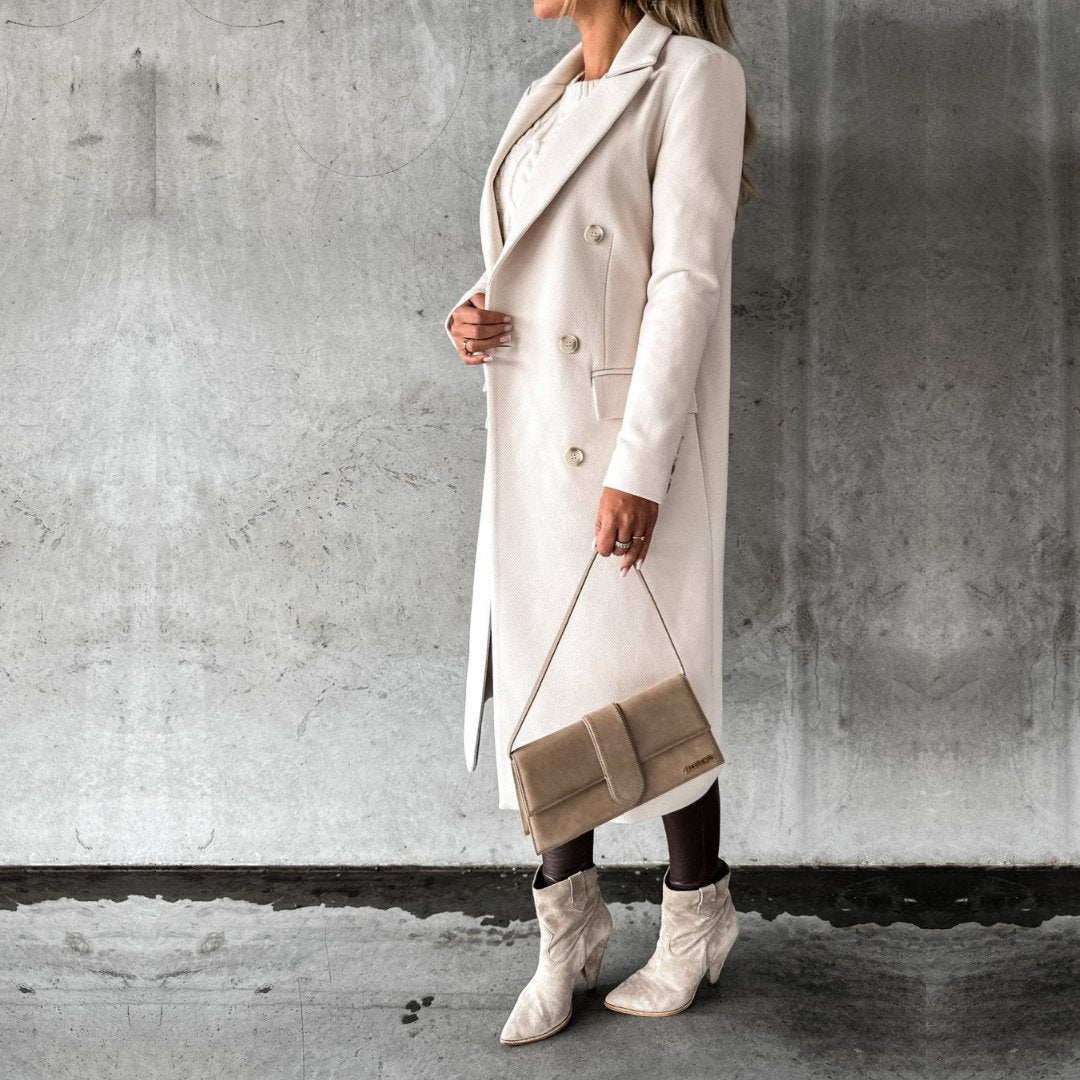Donna in cappotto bianco elegante, stivali alla moda e borsa beige, posa contro parete grigia. Look autunnale chic e sofisticato.