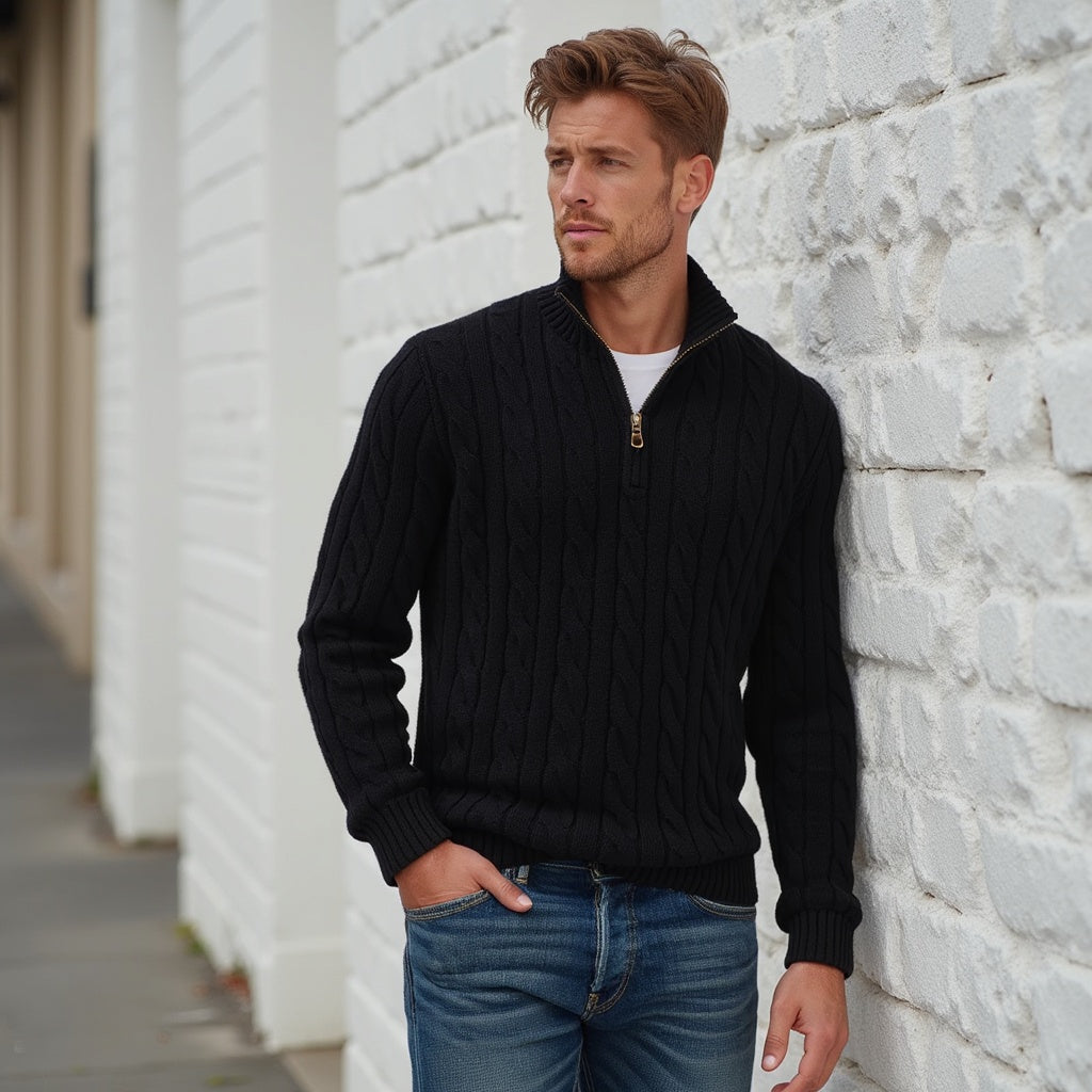 Uomo con maglione nero a trecce e jeans, appoggiato a un muro bianco. Moda maschile autunnale, stile casual elegante.