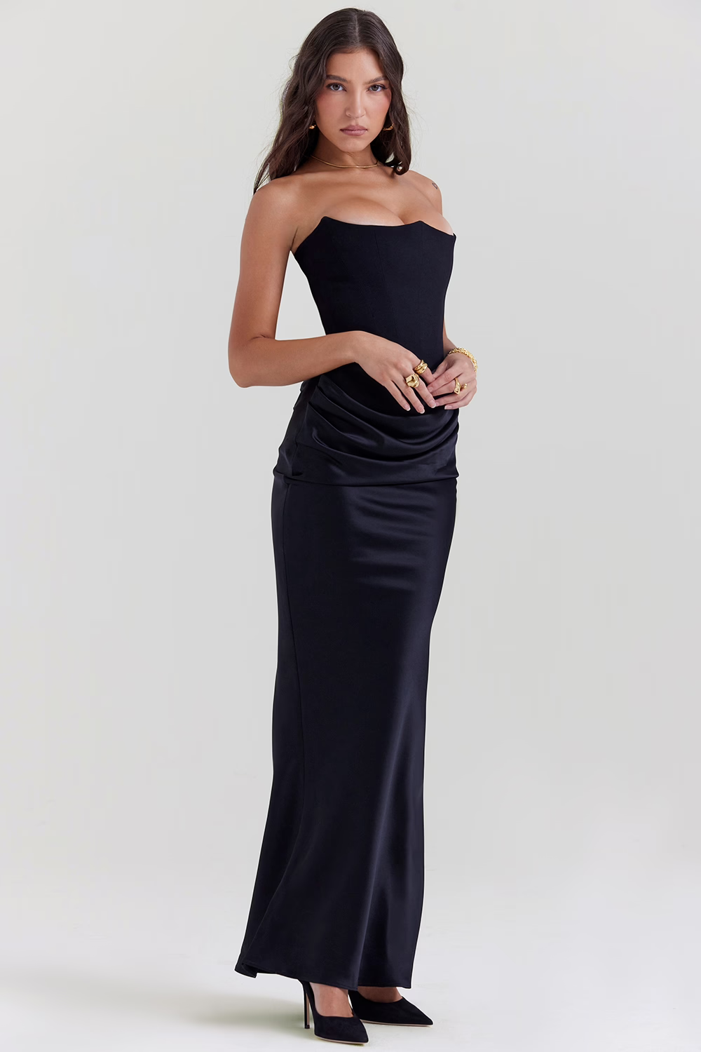 Donna in abito lungo nero elegante, senza spalline, con drappeggio in vita. Indossa tacchi neri e anelli dorati. Moda serale, stile sofisticato.