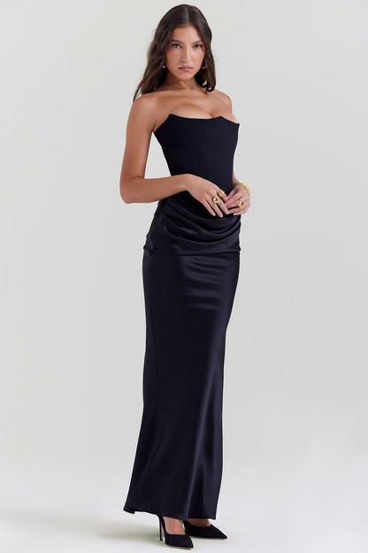Donna in abito lungo nero elegante, senza spalline, con drappeggio in vita. Indossa tacchi neri e anelli dorati. Moda serale, stile sofisticato.