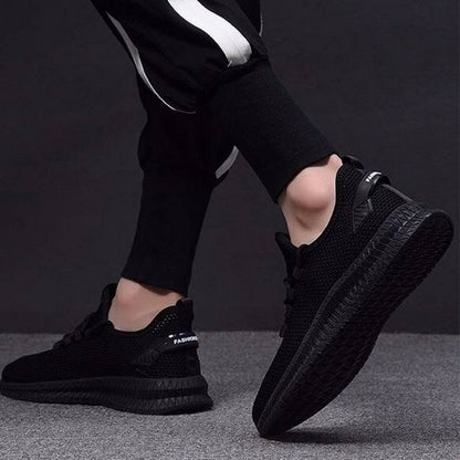 Scarpe da ginnastica nere su sfondo grigio, indossate da una persona in pantaloni sportivi. Design moderno, suola robusta, stile casual.