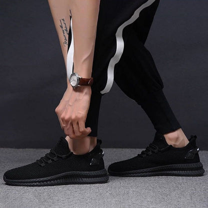 Scarpe da ginnastica nere eleganti, indossate da una persona con pantaloni sportivi neri e orologio al polso, su sfondo grigio scuro.