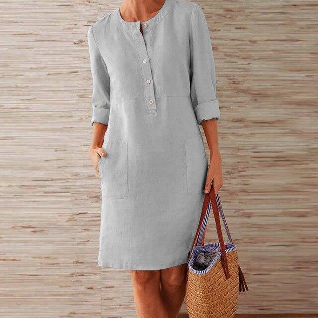 Donna con abito di lino grigio chiaro, maniche lunghe, tasche frontali, tiene una borsa di paglia. Moda estiva casual elegante.