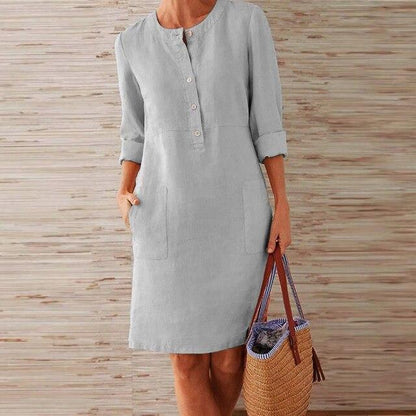 Donna con abito di lino grigio chiaro, maniche lunghe, tasche frontali, tiene una borsa di paglia. Moda estiva casual elegante.