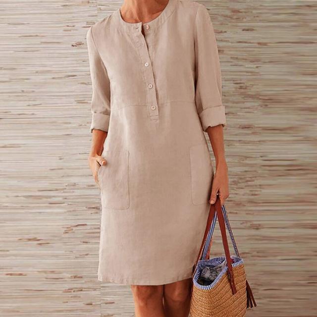 Donna in abito di lino beige con maniche a tre quarti, tasche frontali, accanto a borsa di paglia con manici blu. Moda estiva casual elegante.